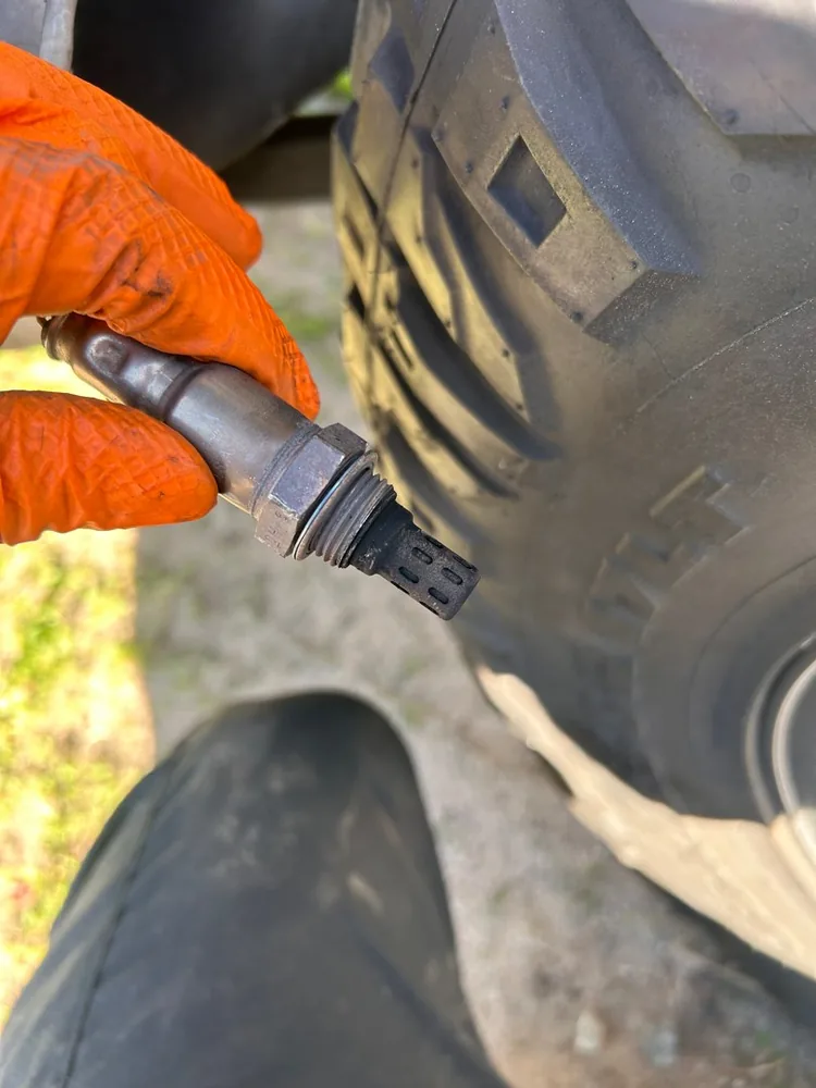 A gloved hand holding an oxygen sensor under a Jeep