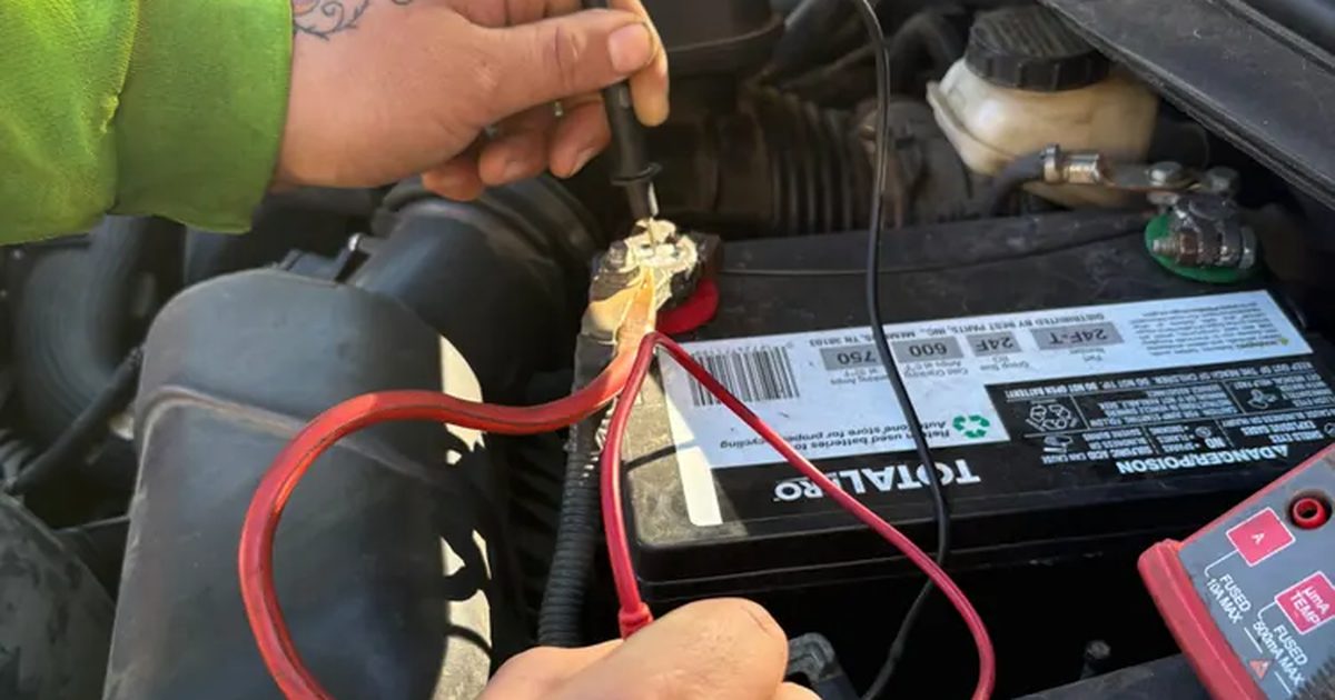 A technician touches multimeter probes to a car battery terminal under the hood