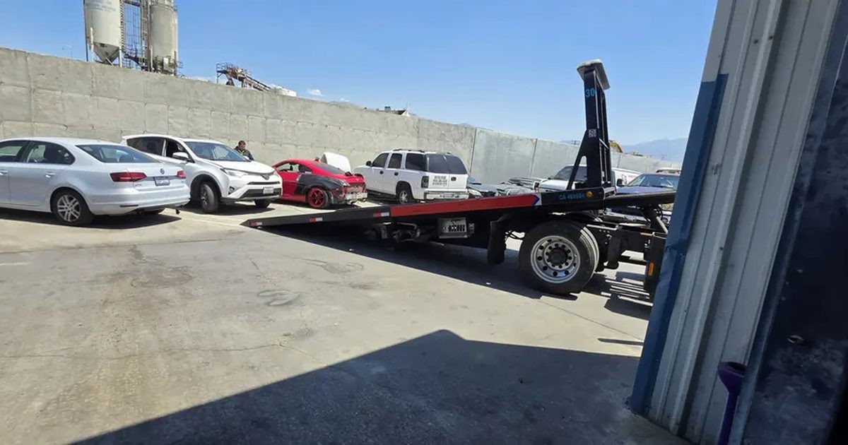 A flatbed tow truck with its bed tilted down, backing up to a row of cars in a shop lot