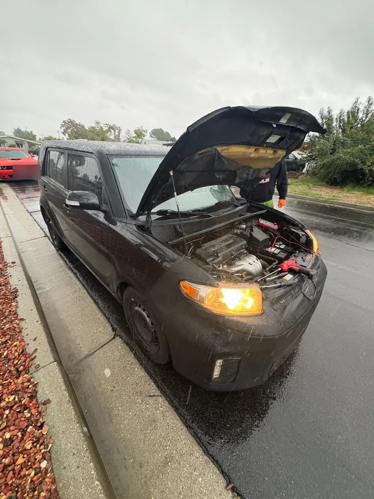 A black Scion with the hood up at the curb in the rain during a roadside jump start