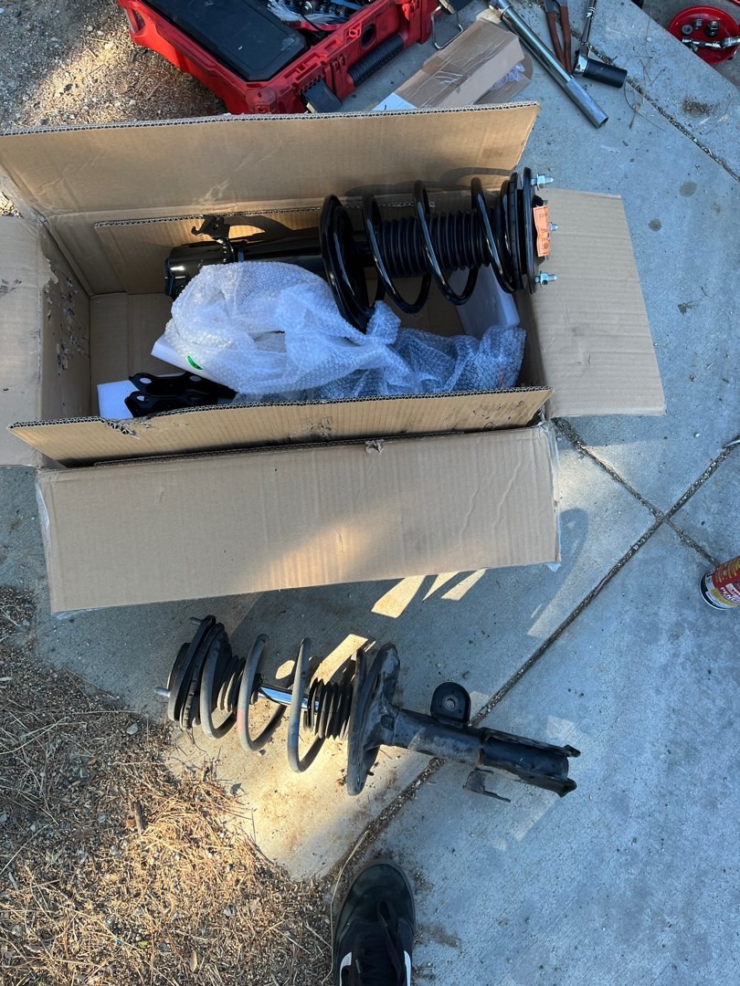 A new loaded strut with its spring in an open box beside the old strut on the driveway
