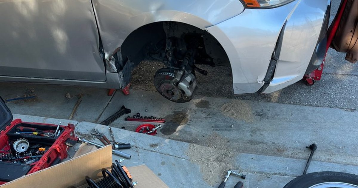 A car jacked up in a driveway with the front wheel and strut off and a new strut in its box