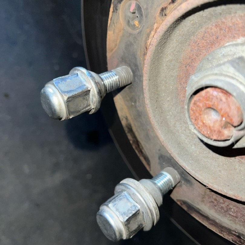 Two lug nuts threaded on wheel studs next to a rusty hub and a rusted axle nut